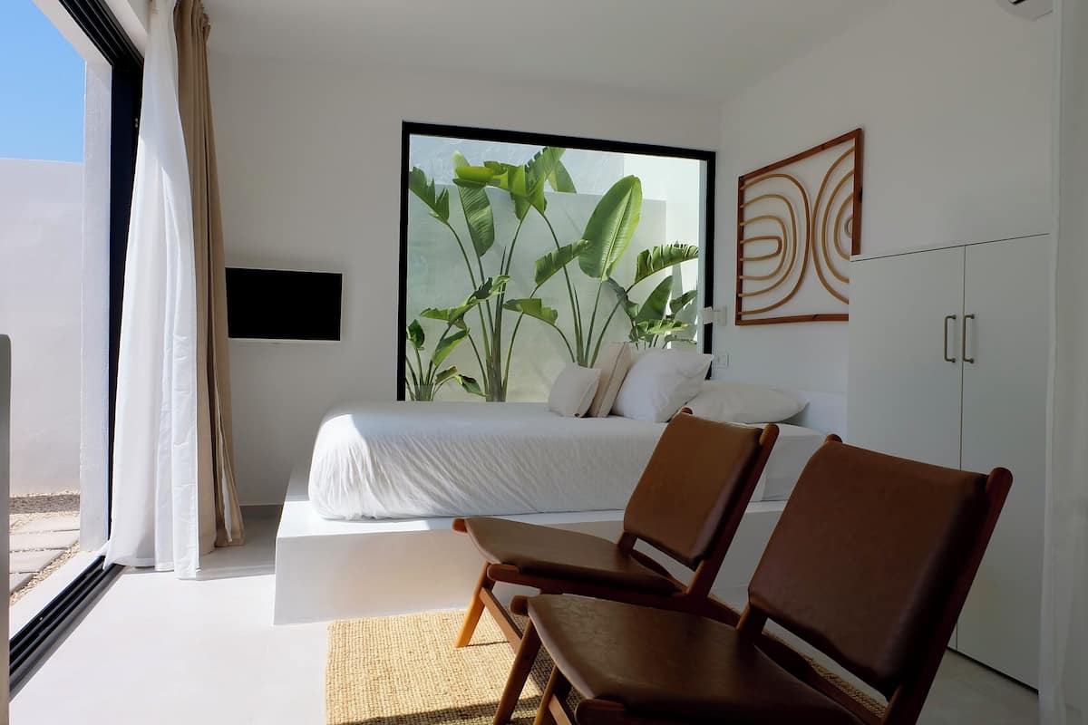 A modern bedroom with a large window, a bed, two wooden chairs, and a wardrobe.