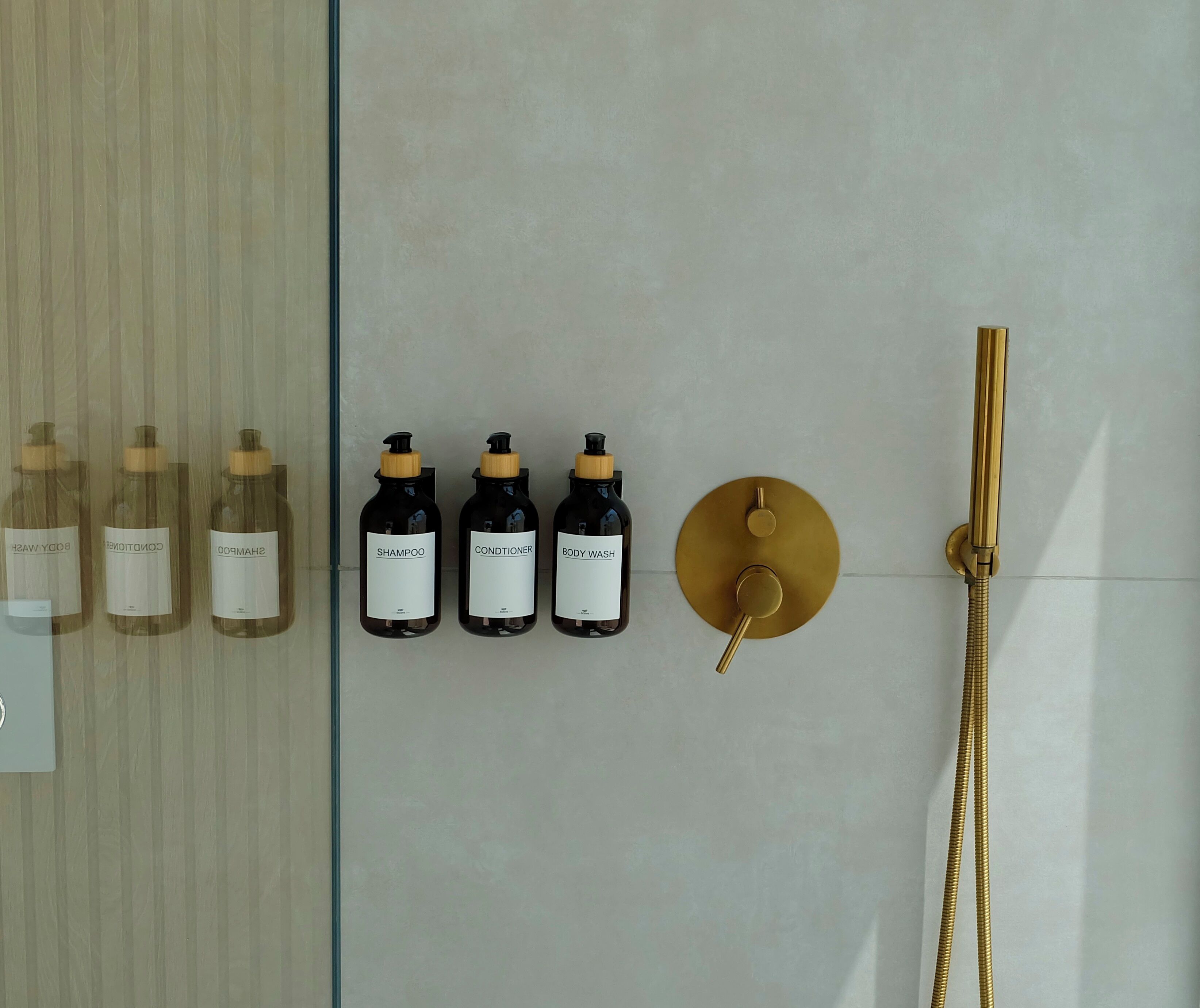 Un baño con mampara de vidrio, ducha con cabezal dorado y botellas de champú, acondicionador y gel de baño.