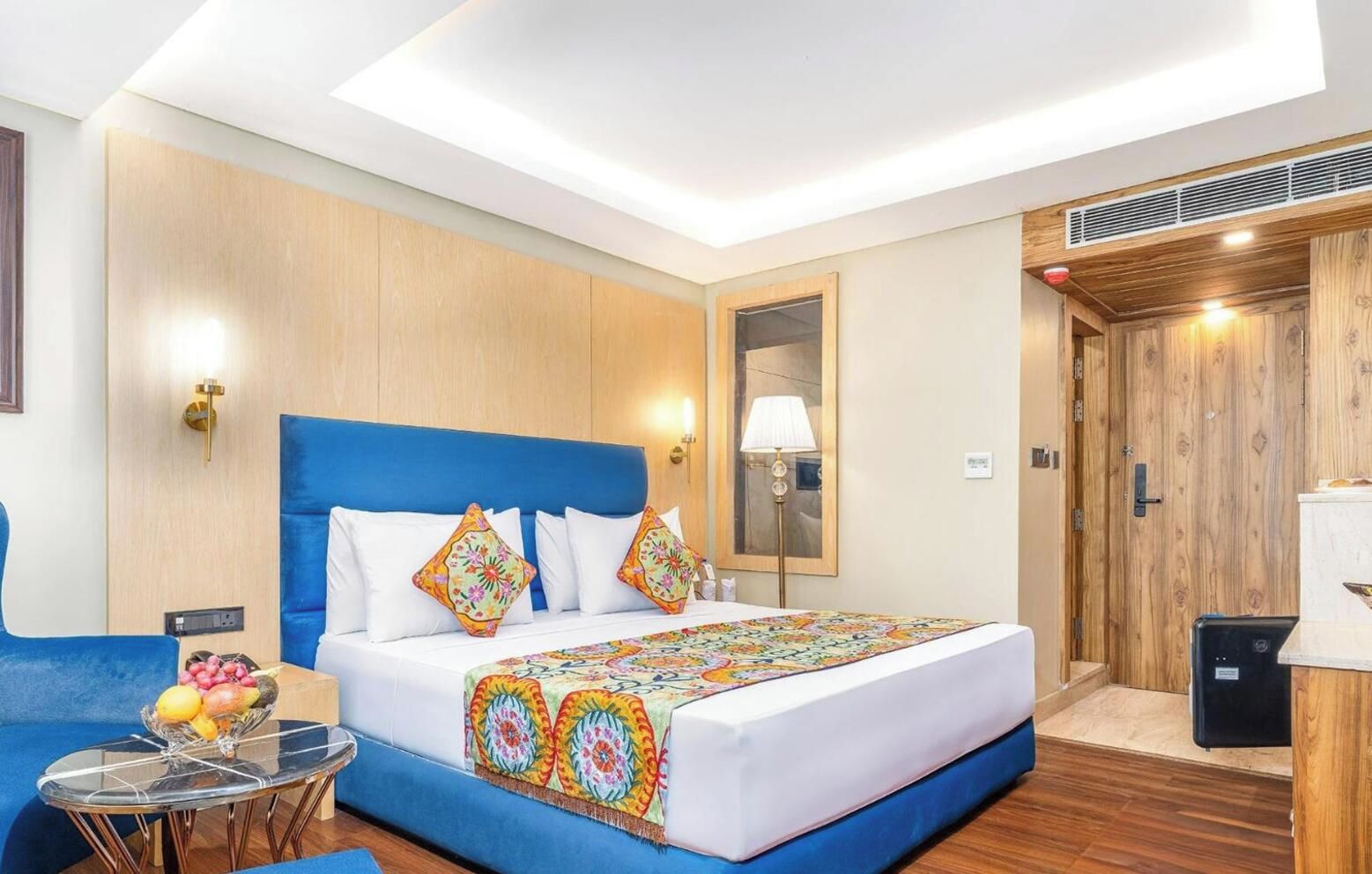 Habitación de hotel con una cama grande, cabecera azul, mesita de noche con frutero, espejo y armario de madera.