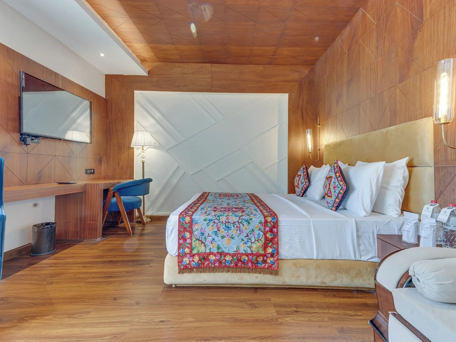 A hotel room with a wooden ceiling, a large bed with colorful pillows, a desk with a chair, and a television mounted on the wall.