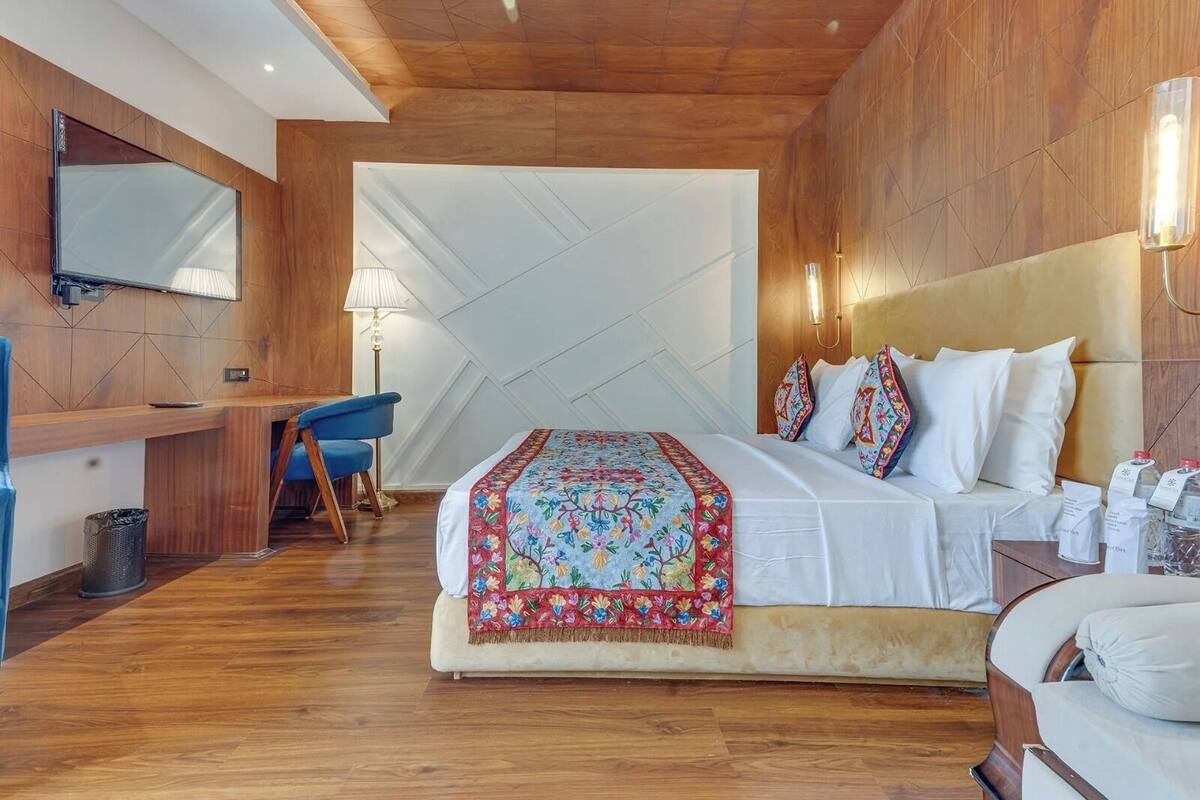 A hotel room with a wooden ceiling, a large bed with colorful pillows, a desk with a chair, and a television mounted on the wall.