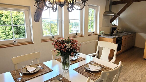 A dining area with a table set for four, a chandelier, and a view of the outdoors through large windows. - Apartment in Winterberg Near Ski Lift (Winterberg)