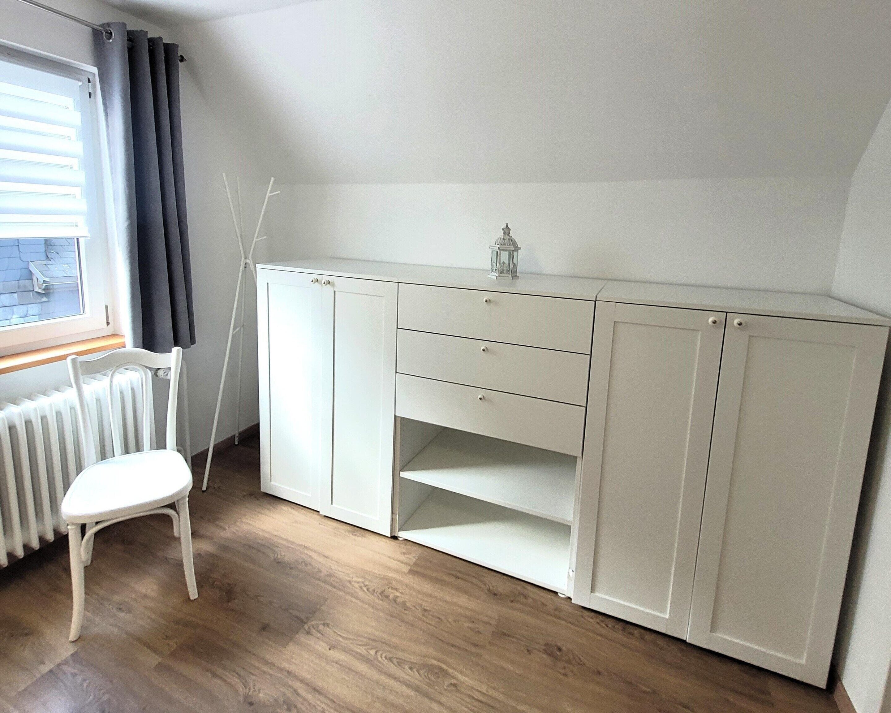 A room with a white wardrobe, a white chair, a radiator, and a window with curtains.