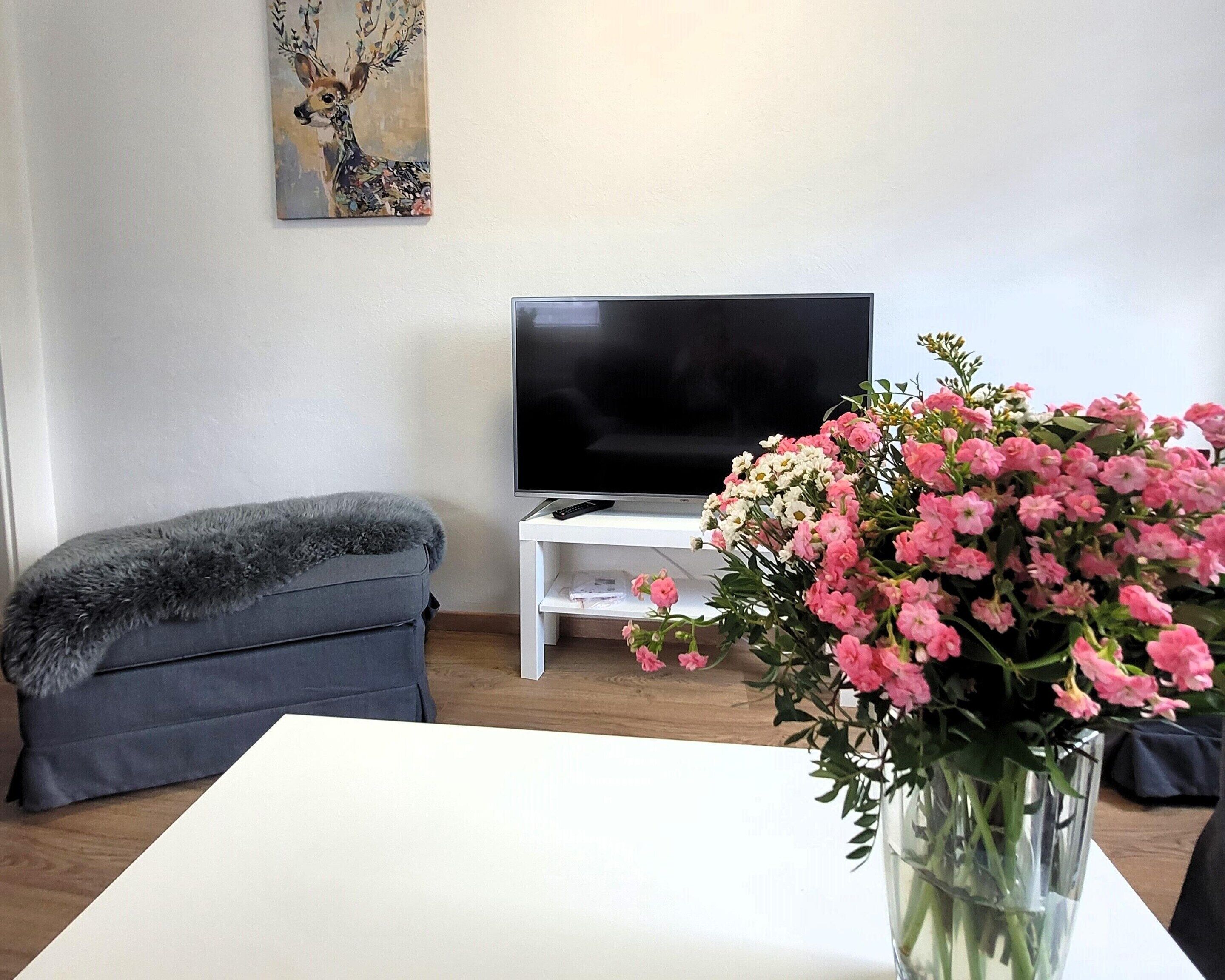 Ein Wohnzimmer mit einem Flachbildfernseher, einem grauen Sofa, einem weißen Tisch und einer Vase mit rosa Blumen.
