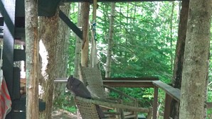 A rustic wooden rocking chair under a covered porch with a view of a forest. -  White Mountain riverside Treehouse (Warren)