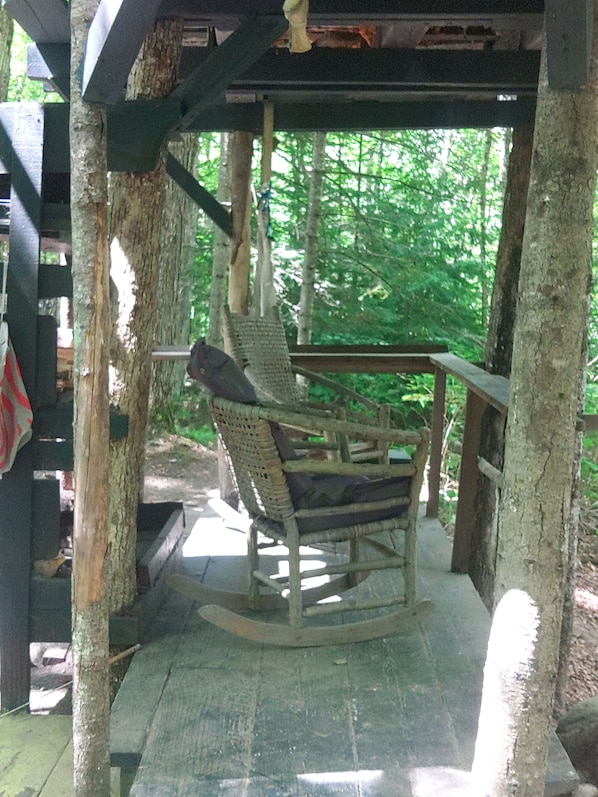 A rustic wooden rocking chair under a covered porch with a view of a forest. - White Mountain riverside Treehouse (Warren)