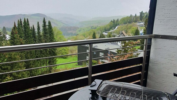 Un balcon doté d’une balustrade en métal, donnant sur une vallée parsemée de maisons et d’arbres.