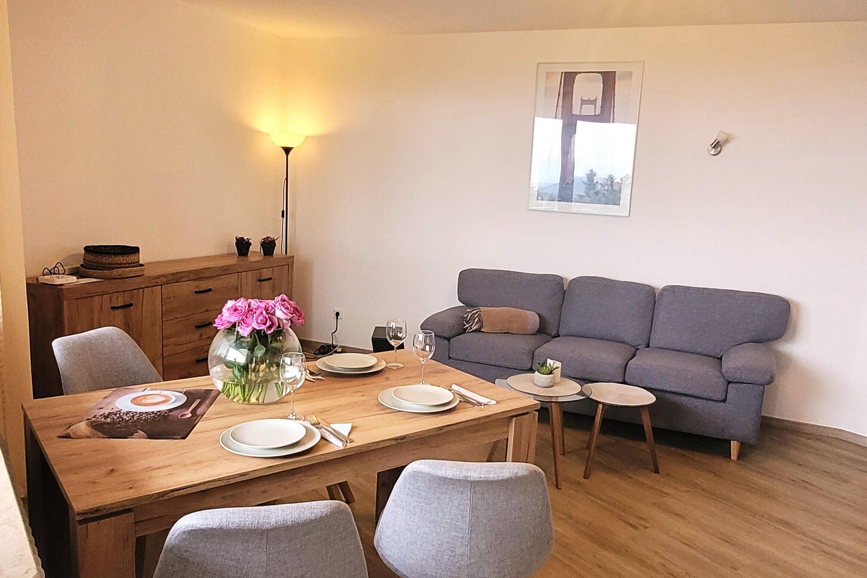 A dining area with a wooden table set for four, a grey sofa, and a small side table with a plant.