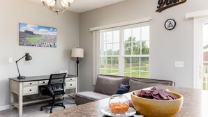 Een kamer met een bureau, een stoel, een bank, een raam met uitzicht op een groen veld, een kroonluchter en een schaal eten op het aanrecht.