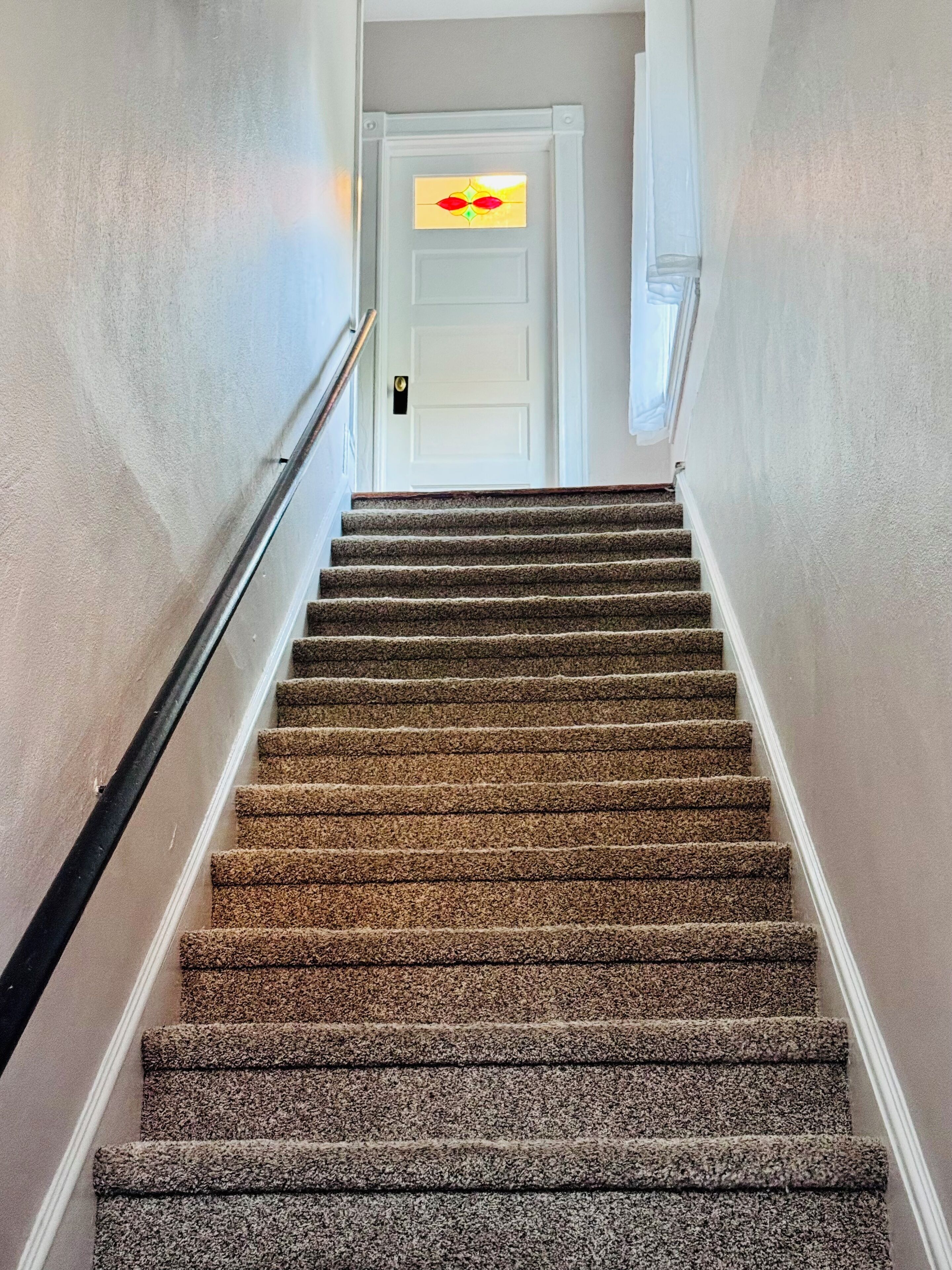 Una escalera alfombrada con barandilla, que conduce a una puerta blanca con un vitral.