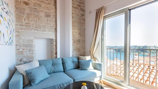 A modern living room with a stone wall, a blue sofa, a black coffee table, and a large window with a view of a cityscape and water. - Seagull's View - Breezy 1 Bedroom with Seaview in the Heart of Rovinj's Old Town (Rovinj)