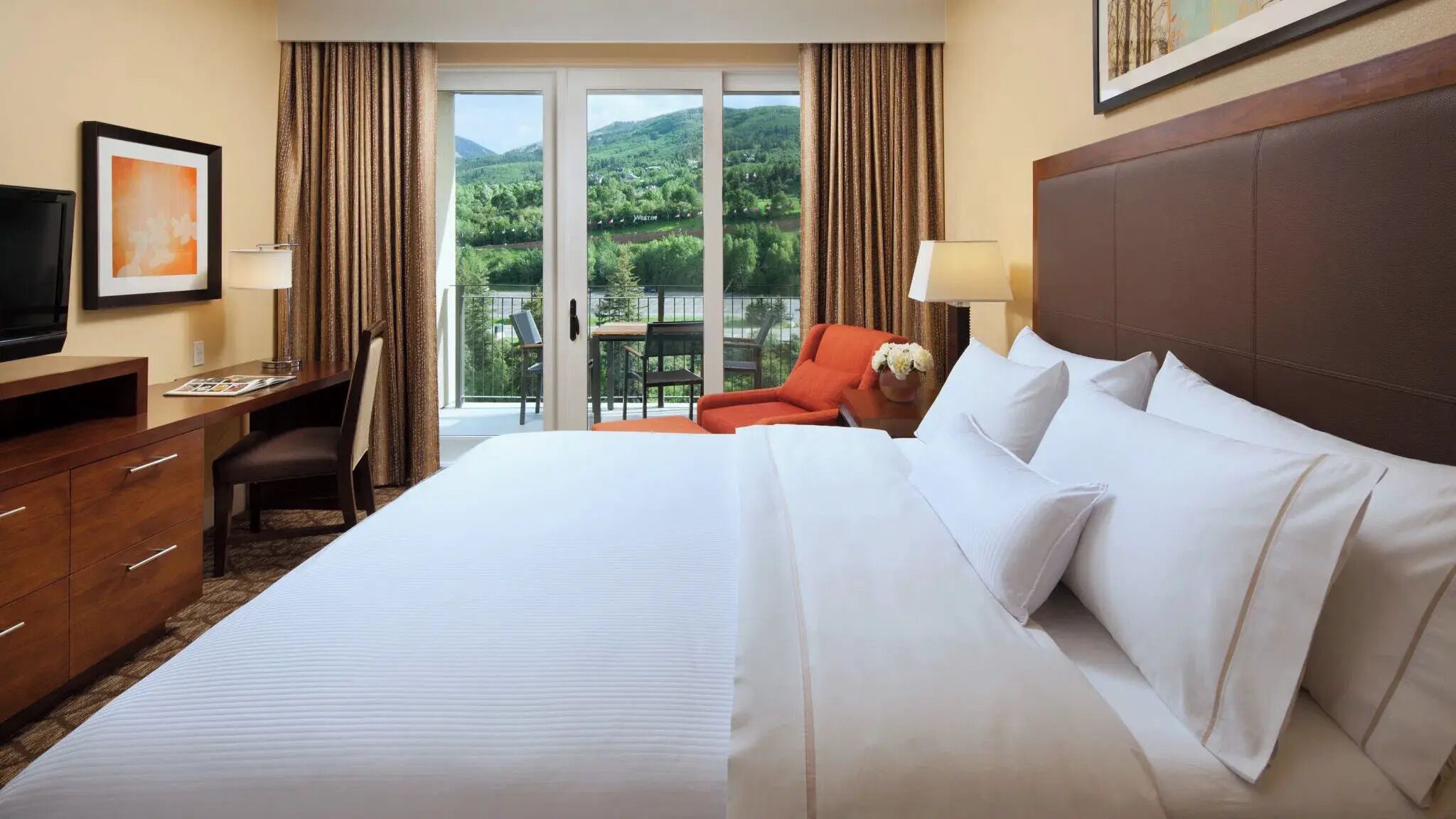 A hotel room with a large bed, a desk with a chair, a television, and a balcony with a view of green hills.