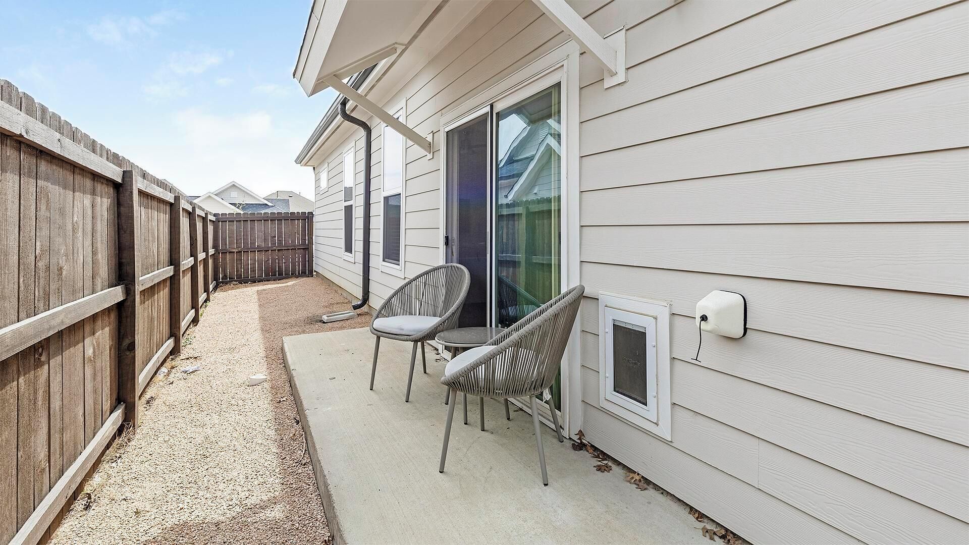 A covered patio with two wicker chairs and a small table, adjacent to a house with white siding and a wooden fence.