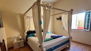 A bedroom with a four-poster bed, a nightstand, a chair, and a window with a view of a building outside. - Rustic Villa with Private Pool near Ocean, Hiking & Cycling Trails – Sleeps 9 (CN)