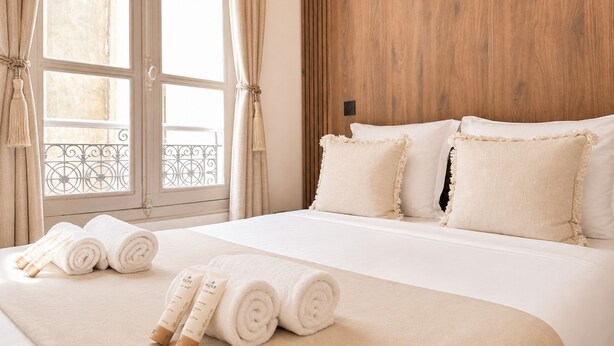 A hotel room with a large bed, two pillows, a wooden headboard, a window with curtains, and a view of a balcony.