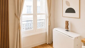 A room with a window, curtains, a radiator, a framed abstract artwork, and a small wooden sculpture on a white table.