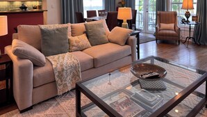A living room with a beige sofa, a glass coffee table, and a kitchen in the background.