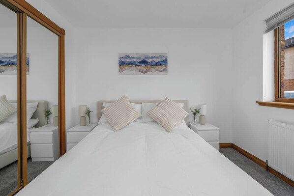 A bedroom with a bed, bedside tables, a mirror, a window, and a painting on the wall.