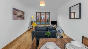A modern living room with a wooden floor, a dark sofa, a dining table set with plates and a glass, a painting on the wall, and a window with a view of a building.