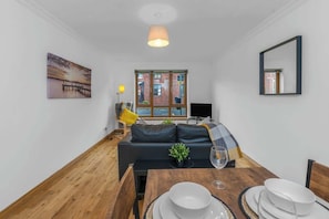 A modern living room with a wooden floor, a dark sofa, a dining table set with plates and a glass, a painting on the wall, and a window with a view of a building.