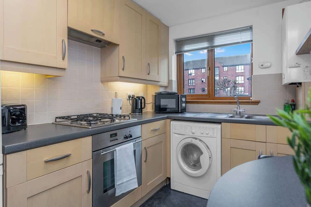 A modern kitchen with a gas stove, microwave, oven, washing machine, and a window with a view of buildings.