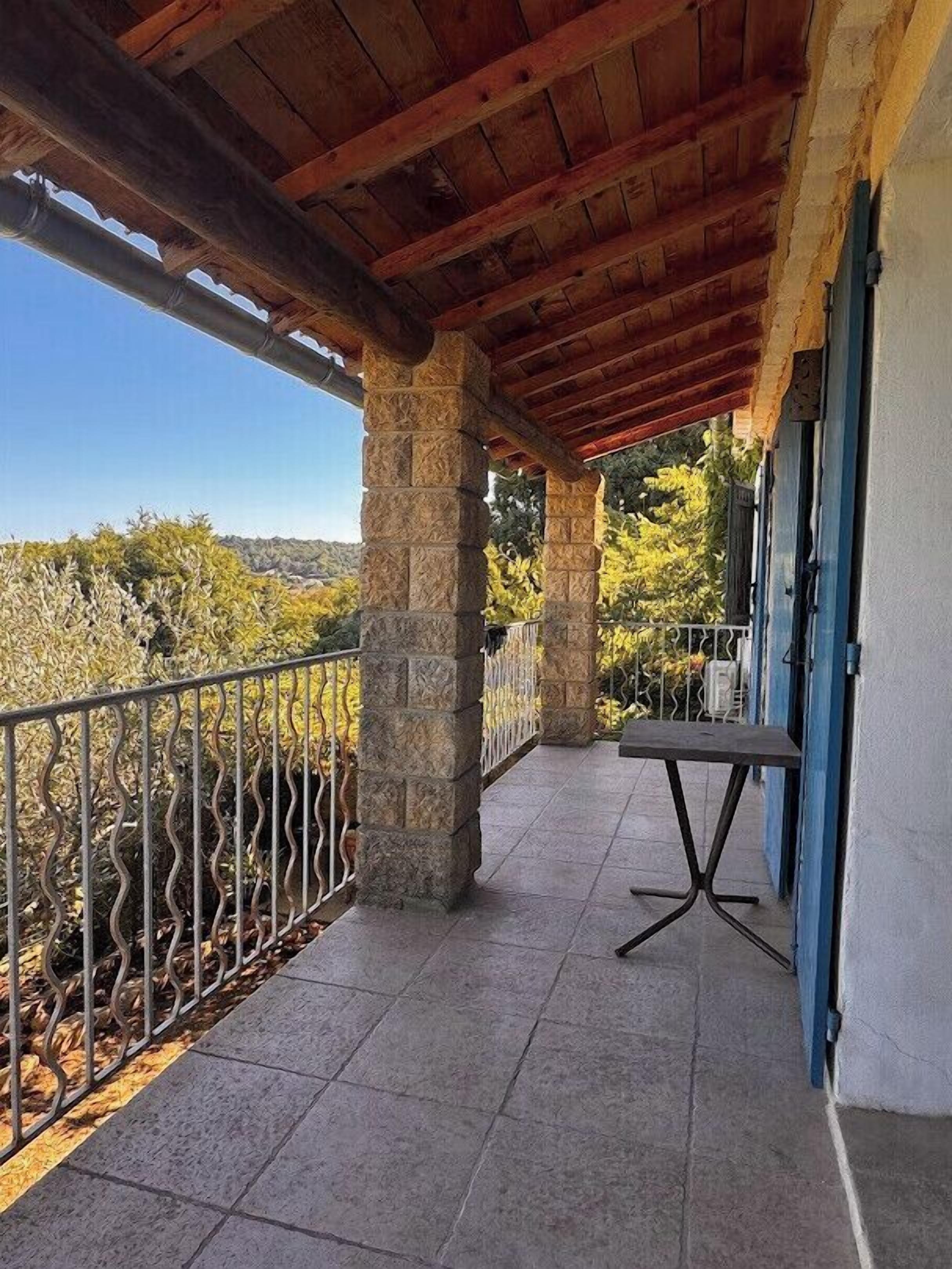 A covered balcony with stone pillars, a metal railing, and a view of trees and hills.