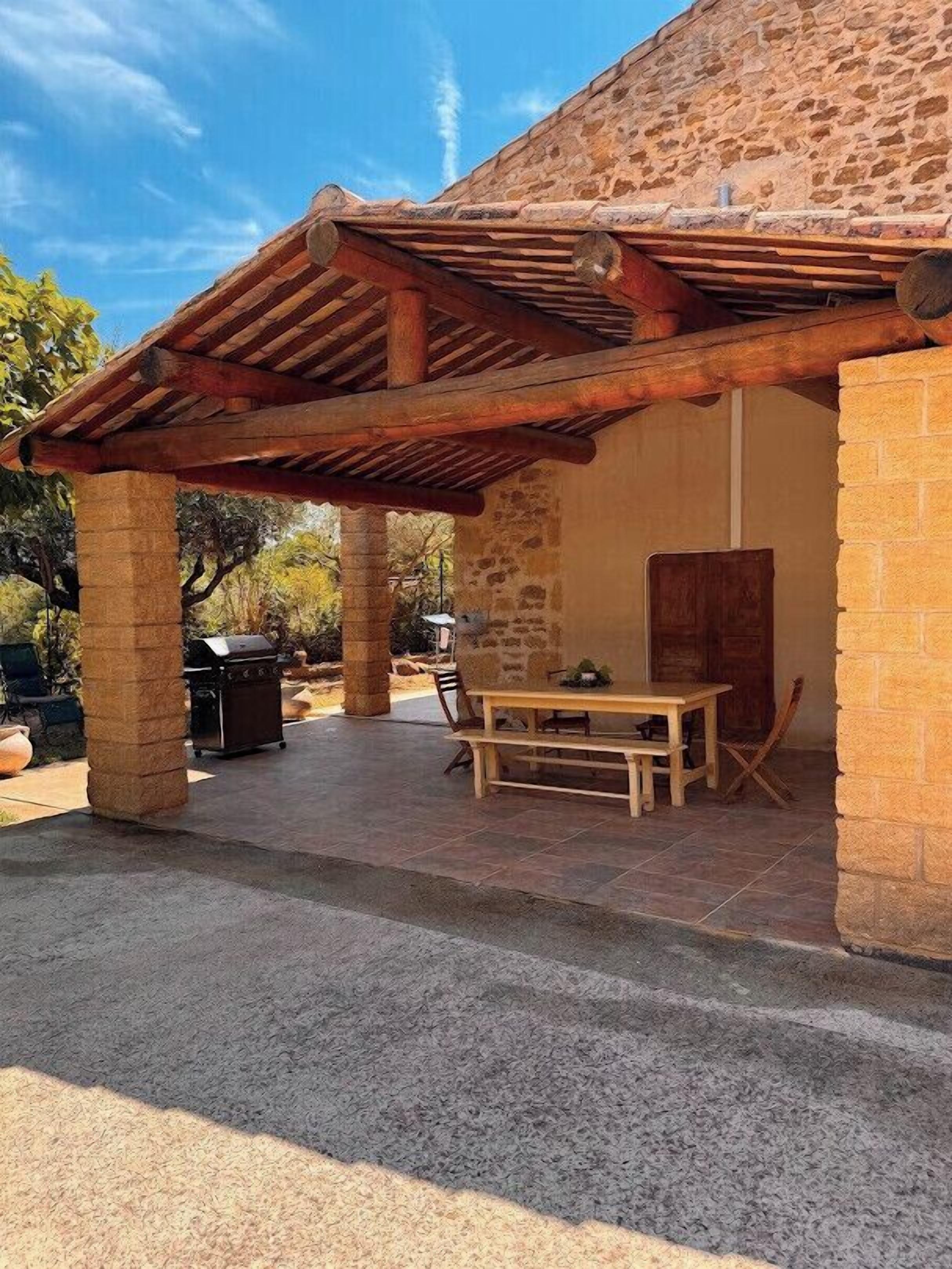 A covered patio area with a wooden pergola, a table with chairs, and a grill.