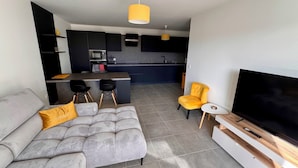 A modern living room with a grey sofa, a yellow armchair, and a TV on a wooden stand. A kitchen with dark cabinets and a yellow pendant light is visible in the background. - Charming 3 rooms located in Le Cannet, near Cannes (Le Cannet)