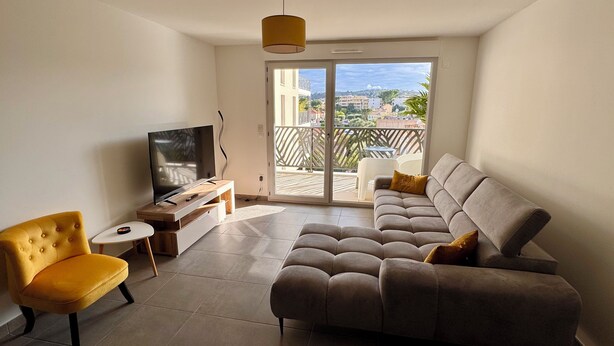 A modern living room with a sofa, a yellow armchair, a TV on a wooden stand, and a balcony with a view of buildings and greenery. - Charming 3 rooms located in Le Cannet, near Cannes (Le Cannet)