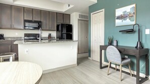 A modern kitchen with dark cabinets, a microwave, stove, and refrigerator. There's a dining area with a round table and a chair. A desk with a chair and a lamp is against a teal wall.