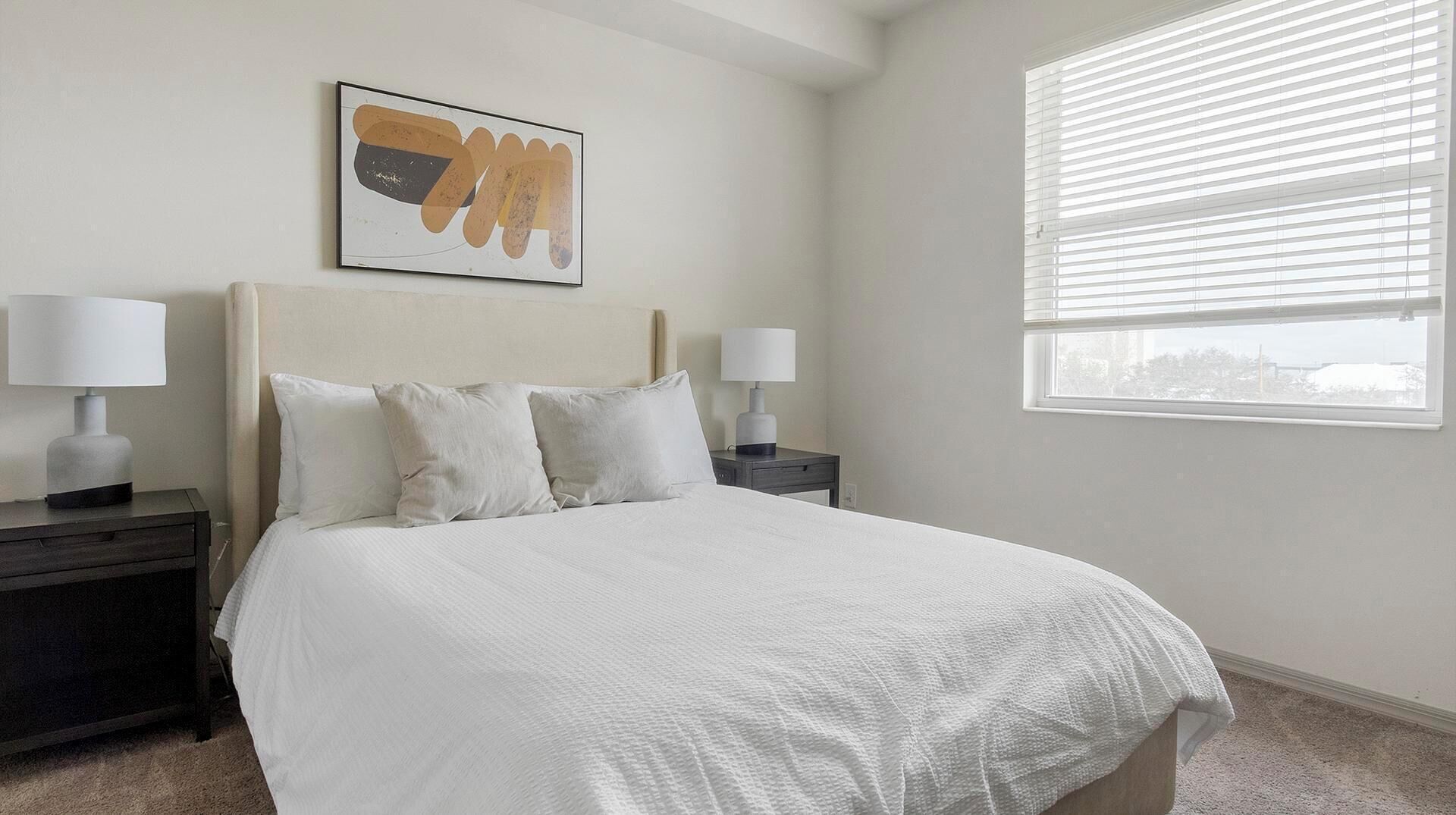 Une chambre à coucher comprenant un lit, deux tables de chevet, une commode, une fenêtre avec des stores et une œuvre d’art accrochée au mur.