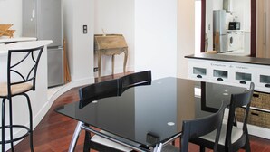 A modern kitchen with a dining area, featuring a glass-top table, black chairs, a wooden side table, and a built-in oven. - Bright apartment with mezzanine in Asnières (Asnières-sur-Seine)