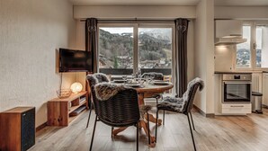Et moderne spiseområde med et træbord og stole, et fjernsyn ophængt på væggen og udsigt til et bjergrigt landskab gennem store vinduer.