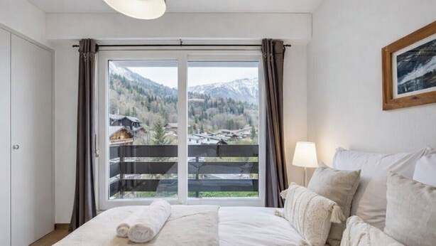 Et soveværelse med seng, et vindue med gardiner, en lampe og et billede i ramme på væggen.