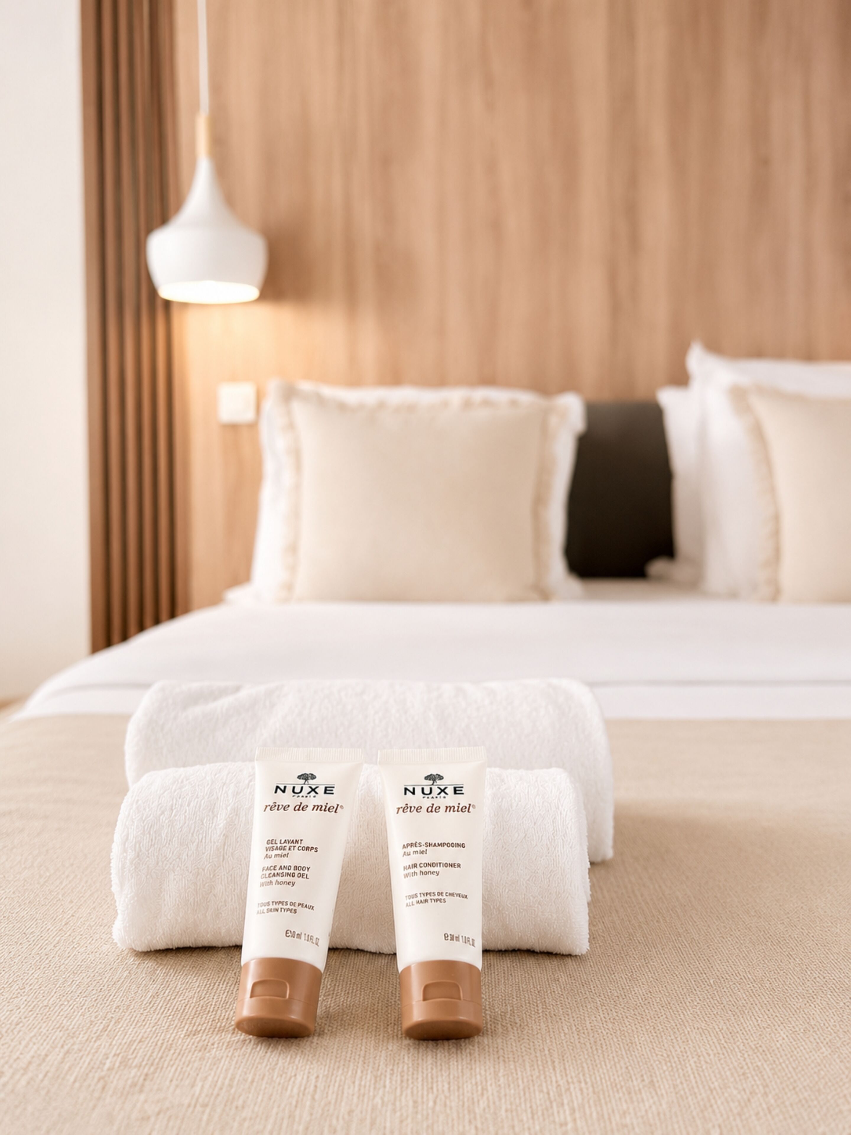 A hotel room with a neatly made bed, a white towel, and two NUXE hair care products on the bed.