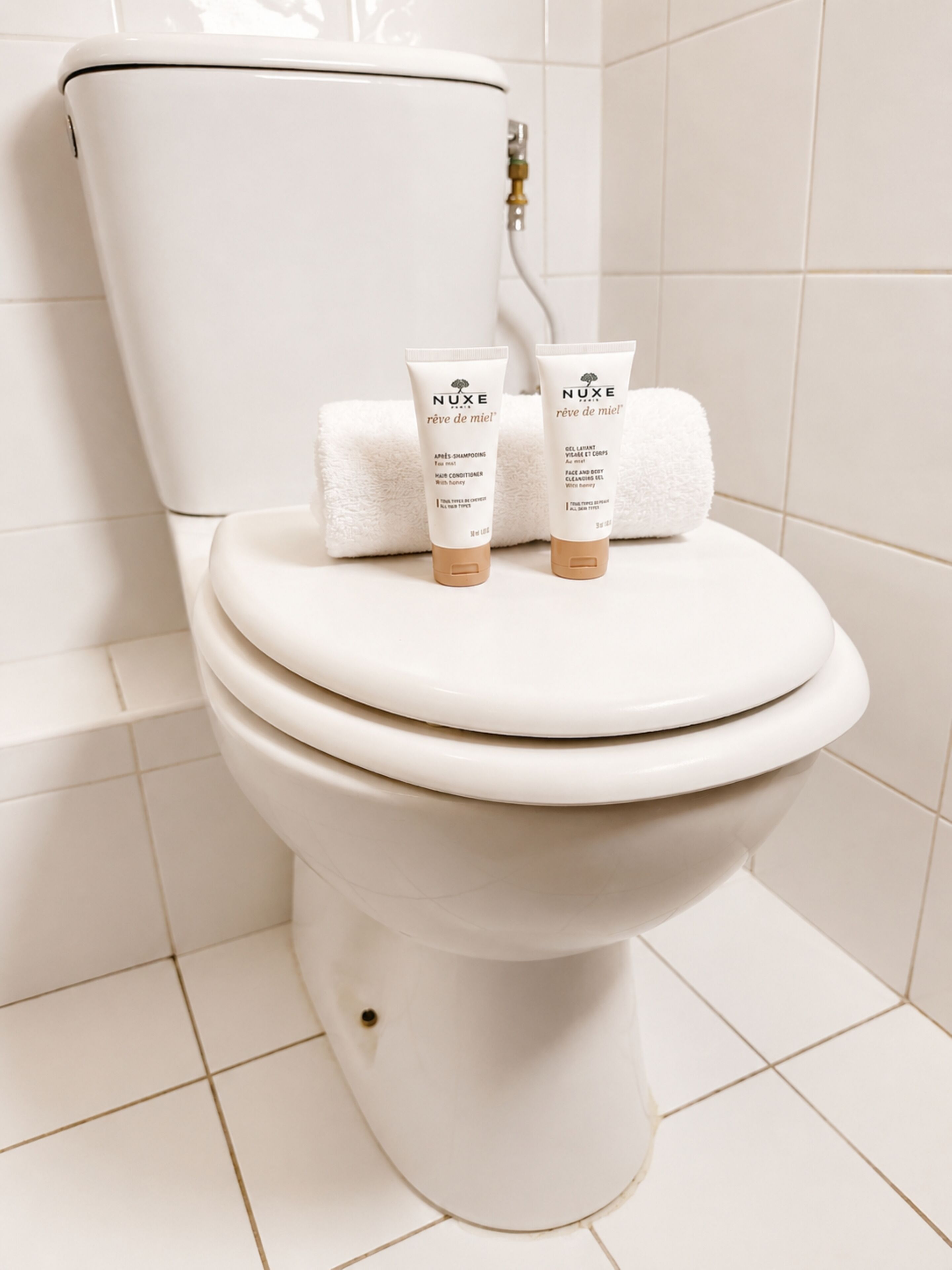 A white toilet with two NUXE shampoo and conditioner tubes and two rolled towels on the lid.