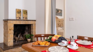 Un comedor con chimenea de piedra, una mesa dispuesta con frutas, pasteles y tazas de café.