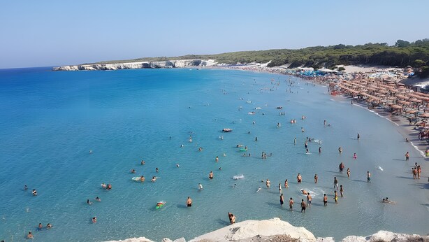 Una playa con agua azul cristalina, gente nadando y tomando el sol, y un litoral rocoso.