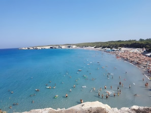 Een strand met helderblauw water, mensen die zwemmen en zonnen, en een rotsachtige kustlijn.