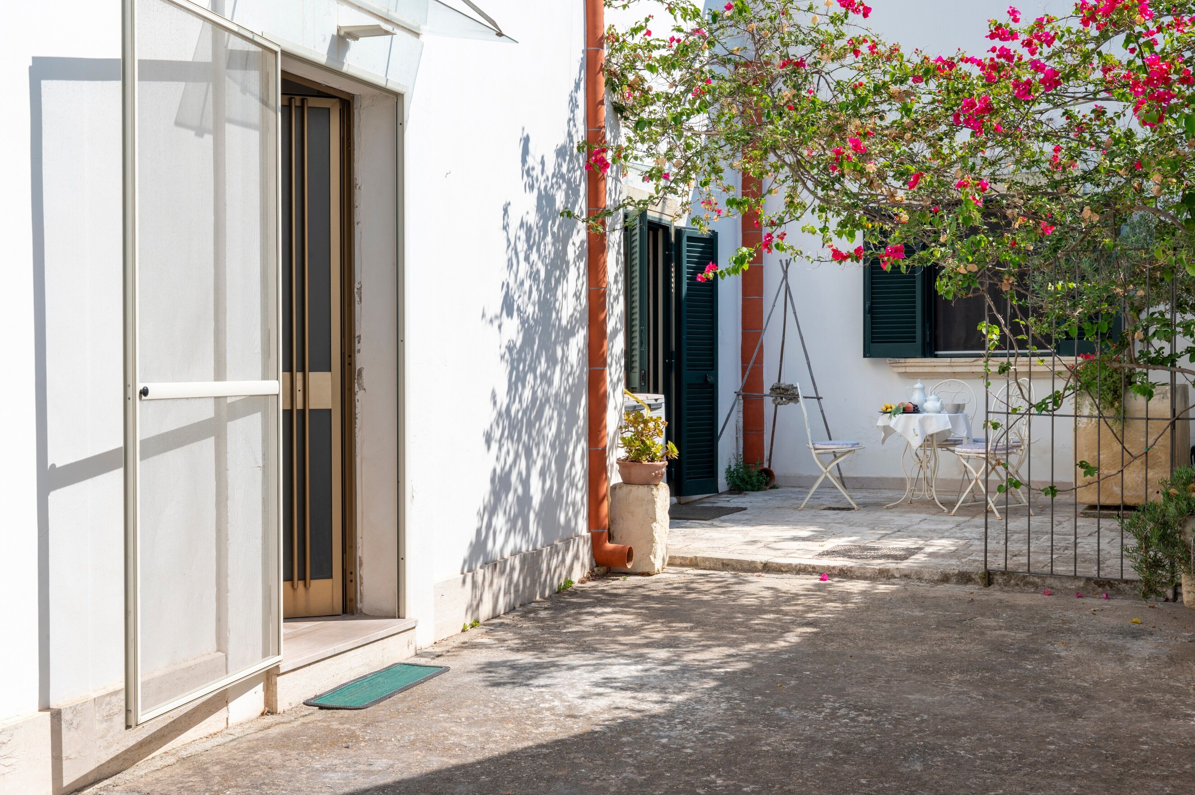 Un cortile con un edificio bianco, una porta, un piccolo tavolo con sedie e una pianta in vaso.