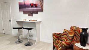 Ett modernt vardagsrum med en vit vägg, en tavla som föreställer en stadsbild, två barstolar, en färgglad fåtölj och en svart vas på ett litet bord i trä.