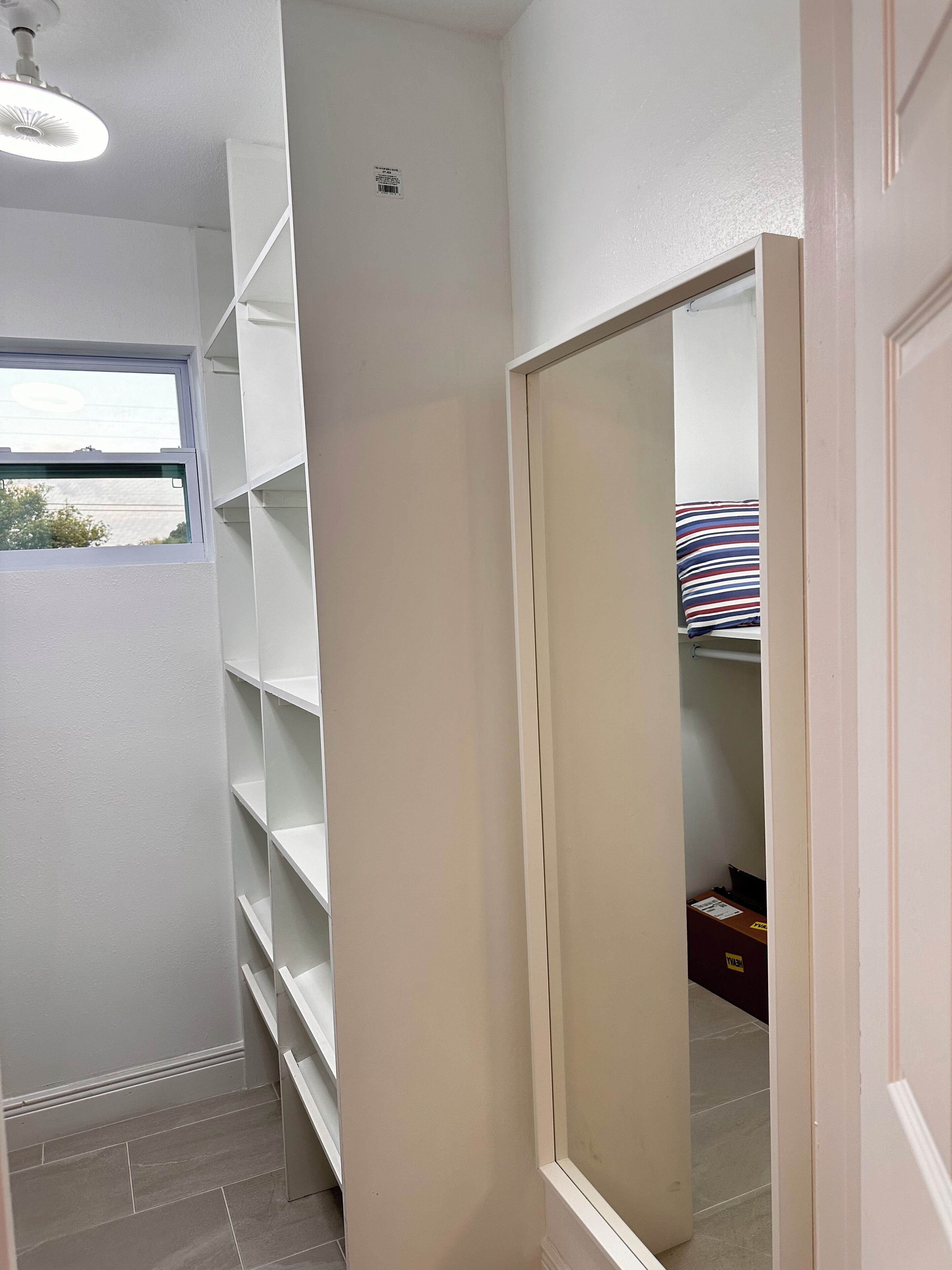 A narrow hallway with white walls, a window, and a door leading to a room with a striped pillow.