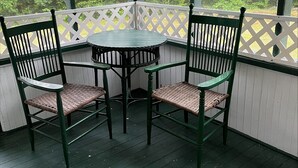A covered outdoor seating area with a table and two chairs, surrounded by a white lattice fence and a view of trees. - Mountain cottage. 4-bdrm cottage in the White Mountains with hiking trails. (Shelburne)