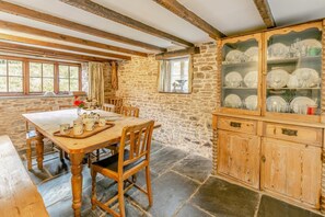 A rustic dining room with a stone wall, wooden beams, a wooden table set with plates and cups, and a glass cabinet with dishes. - 5 Bed in Littleham (oc-b30981) (Littleham)