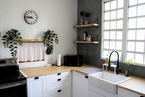 Cozinha com armários brancos, fogão preto, um relógio, duas plantas em vasos, janela com persianas e uma prateleira com garrafas.