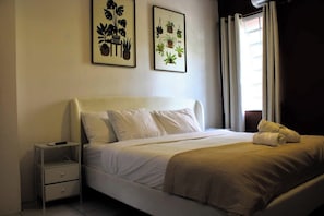 Uma cama bem arrumada com cabeceira branca, uma mesa de cabeceira com abajur e duas gravuras botânicas emolduradas na parede.