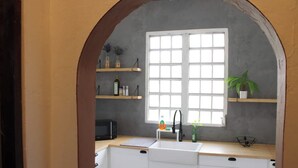 Una cocina con armarios blancos, un microondas negro, un ventanal con contraventanas blancas y estantes con varios objetos.