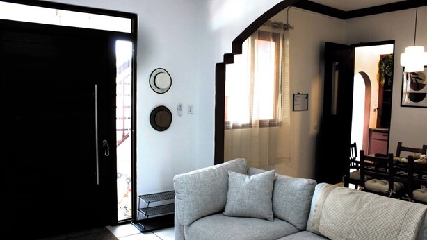 Una sala de estar con sofá, una puerta, una ventana con cortinas y un comedor con sillas.