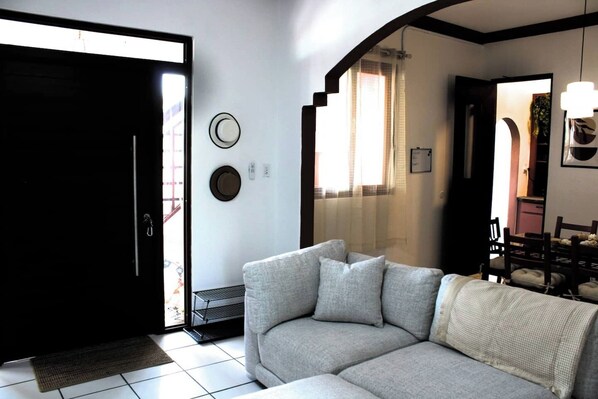 A living room with a sofa, a door, a window with curtains, and a dining area with chairs.