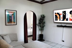 Quarto de hotel moderno com sofá, uma pintura grande, planta em vaso e televisão na parede.